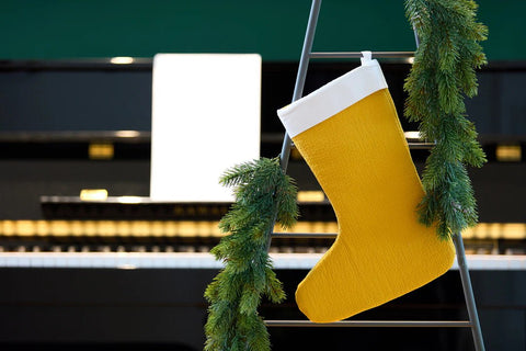 Ein gelber Weihnachtsstrumpf hängt an einer mit Tannengirlanden geschmückten Leiter, die vor einem Klavier mit Notenblättern steht.