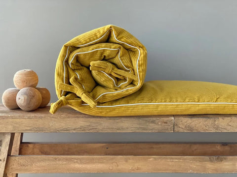 Ein zusammengerolltes senfgelbes Kissen mit Schleifen sitzt auf einer rustikalen Holzbank neben drei Holzkugeln vor einer schlichten grauen Wand.