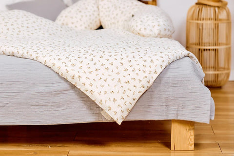 Ein gemütliches Bett mit einem hellgrauen Laken und einer weißen Bettdecke mit kleinem Blumenmuster auf einem Holzrahmen, mit einer Korblampe und Kissen im Hintergrund.