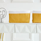 Ein moderner weißer Konsolentisch mit geometrischen Schubladen und goldenen Beinen zeigt zwei KraftKids Körbchen Leinen gelb senfgelb als stilvolle Aufbewahrung und eine minimalistische abstrakte Drahtskulptur vor einer weiß getäfelten Wand.