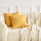 Ein cremefarbenes strukturiertes Sofa mit zwei KraftKids Kissenbezug Leinen gelb senfgelb und einer beigefarbenen strukturierten Decke, die über eine Seite drapiert ist, vor einer weißen Wand mit Zierleisten.