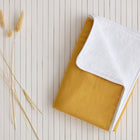 A KraftKids Wickelunterlage Leinen in senfgelb liegt gefaltet mit weißer Umrandung auf einer weißen, gelochten Oberfläche. Links daneben sind dekorativ mehrere getrocknete Gräser arrangiert.