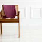 Ein Holzstuhl mit einer Rückenlehne aus geflochtenem Rohr hat einen KraftKids Stillkissenbezug Leinen magenta dunkel über sich drapiert. Der Stuhl steht auf einem hellen Boden vor einer weißen Wand mit dekorativer Vertäfelung.