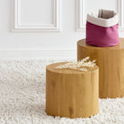 Zwei runde Holzhocker stehen auf einem strukturierten weißen Teppich. Auf dem einen liegen getrocknete Blätter, auf dem anderen das Körbchen rund Leinen magenta dunkel von KraftKids - ideal für die stilvolle Aufbewahrung am Wickeltisch. Dahinter befindet sich eine weiße Wand mit Leisten.