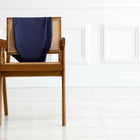 Ein KraftKids Stillkissenbezug Leinen blau dunkelblau (für Theraline 190 cm) aus 100% Leinen liegt über der Rückenlehne eines Holzstuhls mit geflochtener Lehne auf hellem Holzboden vor weißer Wand.