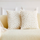 Ein hellgelbes Sofa mit zwei KraftKids Kissenbezug Musselin Zweige braun mit einem kleinen Zweigmuster und einer passenden Decke über der Armlehne. Der Hintergrund zeigt eine weiße Wand mit Zierleisten.