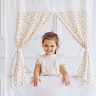 Ein junges Mädchen mit einem rosa Haargummi steht lächelnd vor dem KraftKids Puppentheater Kasperletheater Musselin grau Pusteblumen inkl. Teleskopstange. Teleskopstange und posiert hinter dem zarten, geblümten Vorhang, als wäre es bereit für eine Vorstellung.