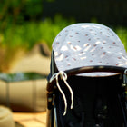 Nahaufnahme eines Kinderwagens mit dem KraftKids Sonnensegel Musselin Kirschen, draußen an einem sonnigen Tag. Unscharfes grünes Blattwerk und beigefarbene Sitzgelegenheiten im Hintergrund betonen den effektiven Sonnenschutz für Babys.