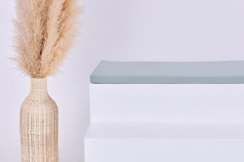 Eine hohe geflochtene Vase mit Pampasgras steht neben einem minimalistischen weißen Podest, das mit der KraftKids GmbH Sitzauflage für Ikea TROFAST Musselin mint gekrönt ist, alles vor einem schlichten weißen Hintergrund.