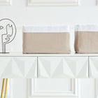 Ein moderner weißer Konsolentisch mit goldenen Beinen bietet Platz für zwei KraftKids Körbchen Musselin beige und eine minimalistische abstrakte Metallskulptur, die vor einer weiß getäfelten Wand steht.