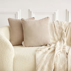 Ein cremefarbenes Struktursofa mit zwei beigen KraftKids Kissenbezug Musselin und einer weichen, gemusterten cremefarbenen Decke, die über die Seite drapiert ist, vor einer weißen Wand mit Leisten - perfekt, um jede Bettwäschesammlung zu ergänzen.