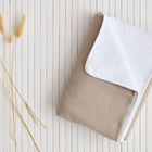 Ein beigefarbenes Falthandtuch mit weißer Ecke liegt auf einer weißen Lattenunterlage neben getrockneten Weizenhalmen, daneben die KraftKids Wickelunterlage Musselin beige für zusätzlichen Komfort.