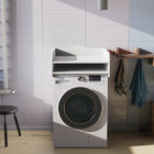 Eine moderne Waschküche mit einem KraftKids Wickelaufsatz weiß für Waschmaschine oder Trockner mit doppeltem Boden, Regalen für Stauraum Wickelutensilien, zwei Handtüchern auf Wäscheklammern, einem runden Spiegel, einem schwarzen Hocker und Sonnenlicht durch Jalousien.