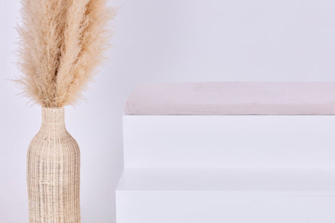 Eine große geflochtene Vase mit Pampasgras steht neben einem minimalistischen weißen Stufenpodest, gekrönt von der KraftKids GmbH Sitzauflage für Ikea TROFAST Cord Breitcord Beige Taupe, vor einem schlichten hellen Hintergrund.