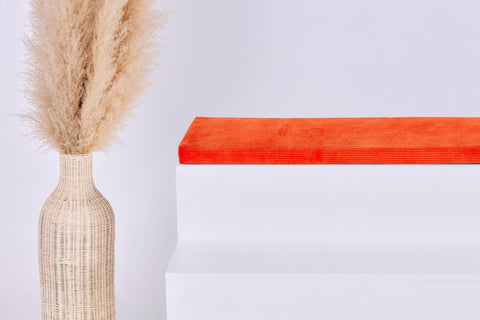 Eine hohe geflochtene Vase mit Pampasgras steht neben einem weißen Stufenpodest, auf dem die KraftKids GmbH Sitzauflage für Ikea TROFAST Cord Breitcord Orange Reinorange steht, alles vor einem schlichten weißen Hintergrund.