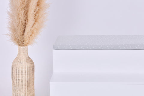 Eine hohe Korbvase mit beigem Pampasgras steht neben einer weißen, minimalistischen Bank, auf der die KraftKids GmbH Sitzauflage für Ikea TROFAST Musselin grau Pusteblumen steht. Der Hintergrund ist einfarbig weiß.