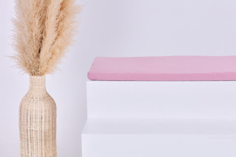 Eine Sitzauflage für Ikea STUVA/SMÅSTAD Musselin rosa von KraftKids GmbH sitzt auf einer weißen Fläche neben einer Vase mit getrocknetem Gras und setzt einen gemütlichen Akzent in Ihrem Raum.