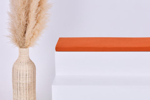 Eine hohe geflochtene Vase mit Pampasgras steht neben einem weißen Stufenpodest, auf dem das KraftKids GmbH Sitzauflage für Ikea TROFAST Doppelkrepp Rot Herbstrot Kissen liegt, alles vor einem schlichten hellen Hintergrund.