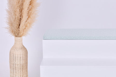 Eine hohe, geflochtene Vase mit beigem Pampasgras steht neben einer minimalistischen weißen Bank mit dem Kissen KraftKids GmbH Sitzauflage für Ikea TROFAST Musselin mint Pusteblumen. Der Hintergrund ist schlicht und hell gehalten.