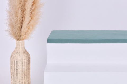 Eine hohe geflochtene Vase mit Pampasgras steht neben einer minimalistischen weißen Bank mit einem KraftKids GmbH Sitzauflage für Ikea TROFAST Musselin nilblau Kissen vor einem schlichten hellen Hintergrund.