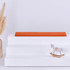 Ein weißes, dreistufiges Display mit einer roten KraftKids GmbH Sitzauflage für IKEA TROFAST Doppelkrepp Herbstrot auf der obersten Stufe, einer Vase mit beigem Pampasgras auf der linken Seite und einem kleinen Holzschaukelpferd auf der rechten Seite vor einem hellen Hintergrund.