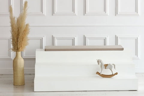 Ein minimalistisches Interieur mit weiß getäfelten Wänden, einer weißen Stufenbank mit der KraftKids GmbH Sitzauflage für Ikea TROFAST Musselin beige, einem dekorativen Holzschaukelpferd und einer hohen geflochtenen Vase mit getrocknetem Pampasgras.