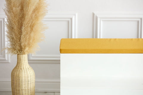 Eine hohe geflochtene Vase mit Pampasgras steht neben einer weißen Bank mit der KraftKids GmbH Sitzauflage für Ikea TROFAST Leinen in senfgelb, die an einer weiß getäfelten Wand steht.