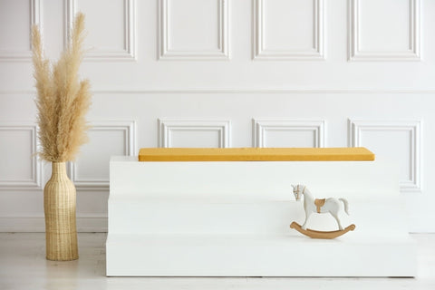Ein minimalistischer Raum mit einer weiß getäfelten Wand, einer hohen Vase mit Pampasgras, weißen Sitzstufen mit einer KraftKids GmbH Sitzauflage für Ikea TROFAST Leinen in gelb senfgelb und einem kleinen Holzschaukelpferd auf der untersten Stufe.