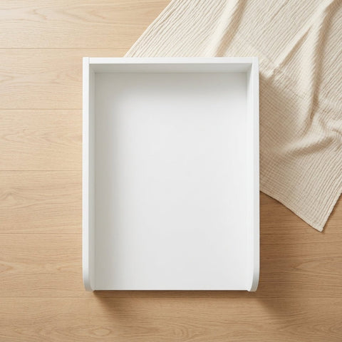 Ein weißer KraftKids Wickelaufsatz universal mit durchgehendem Boden (Wickelfläche 50 x 70 cm) steht leer und rechteckig auf hellem Holzfußboden; oben rechts ist beigefarbener Stoff teilweise sichtbar.