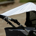 Eine Nahaufnahme eines Kinderwagens im Freien, der mit dem KraftKids Sonnensegel graue unregelmäßige Punkte auf Weiß über dem Verdeck als Schattenspender drapiert ist. Das weiße Sonnensegel weist unregelmäßige graue Punkte auf, der Hintergrund ist unscharf.