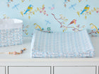 Ein KraftKids-Wickeltisch mit dem "Bezug für Keilwickelauflage kleine Dreiecke blau grau weiß", einer geometrischen blau-grau-weißen Wickelauflage, einem Aufbewahrungskorb und einer Schnullerklemme, vor einer Tapete mit bunten Vögeln und Libellen.