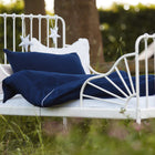 Ein weißes Metallbett mit KraftKids Bettwäscheset Musselin dunkelblau wird im Freien auf Gras aufgestellt. Am Rahmen hängt Sternendekor, wodurch es ideal für erholsamer Schlaf umgeben von Bäumen und üppigem Grün ist.