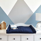 Eine weiße Kommode mit einer KraftKids Wickelauflage Musselin dunkelblau, Babykleidung und einer Haarbürste obenauf. Dekorative Akzente setzen eine Lampe, ein Plüschhase, ein Hausregal mit einem ausgestopften Lamm und geometrische Wandkunst für ein gemütliches Ambiente.