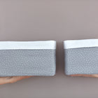 Zwei Hände halten zwei KraftKids Körbchen Musselin grau Punkte vor einer neutralen Wand hoch. Perfekt zur Aufbewahrung am Wickeltisch: Die grauen Stoffkörbe mit weißen Punkten und Band oben kommen im 2er-Set, ein größeres, ein kleineres.