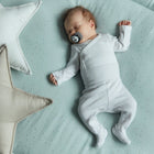 Ein Baby im weißen Schlafanzug schläft friedlich auf einer grünen KraftKids Krabbeldecke Musselin mit goldenen Punkten und weicher Polyestervlies-Füllung, in der Hand einen Schnuller. Große, weiche, sternförmige Kissen sind in der Nähe platziert.