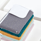 Ein sauber gefalteter Stapel der KraftKids Wickelunterlage Musselin goldene Punkte auf Grau, mit weißen Eckdetails. Die obere graue Decke mit goldenen Punkten ist ideal als wasserundurchlässige Unterlage oder Wickelauflage Baby.