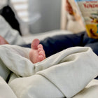 Der nackte Fuß eines Babys lugt unter dem weichen KraftKids Bettwäscheset Leinen in leicht grünem Weiß hervor, während ein Erwachsener im Hintergrund ein aufgeschlagenes Kinderbuch hält. Die gemütliche Umgebung ist perfekt für erholsamen Schlaf.