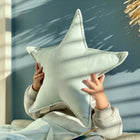 Ein Kind sitzt an einer blauen Wand in einem gemütlichen Kinderzimmer und hält sich das KraftKids Sternkissen Leinen Natur Braun vor das Gesicht, während das Sonnenlicht sanfte Schatten auf die Szene wirft.