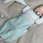 Ein Baby schläft friedlich in einem beigen Bettchen, eingewickelt in den grünen KraftKids Strampelsack Frühling Sommer Musselin mit goldenen Punkten und mit einem Schnuller in der Hand. Ein sternförmiges Kissen liegt daneben und schafft die perfekte Kulisse für erholsame Träume.
