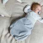 Ein Baby liegt mit einem Schnuller auf einem beigen Bett, trägt ein weißes Oberteil und den KraftKids Strampelsack Frühling Sommer Musselin grau Punkte. Das Baby blickt nach oben, in der Nähe liegt ein großes sternförmiges Kissen.