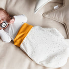 Ein schlafendes Baby mit einem Schnuller liegt auf einem beigen Bett, eingehüllt in den kuscheligen KraftKids Strampelsack Herbst Winter Musselin goldene Punkte auf Weiß. Weiche Kissen, darunter ein sternförmiges, liegen in der Nähe.