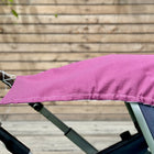 Ein KraftKids Sonnensegel Musselin purpur ist über den Griff und die Seite eines Kinderwagens mit grauem Rahmen drapiert. Im Hintergrund sind ein Holzlattenzaun und ein Teil eines Holzdecks zu sehen.