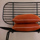 Zwei Kissenbezüge Doppelkrepp Rot Herbstrot von KraftKids sind auf einem modernen schwarzen Metallstuhl mit Lattenrost vor einer hellbeigen Wand gestapelt.