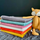 Ein Stapel gefalteter KraftKids Wickelunterlage Cord Breitcord Grau ruht auf grauem Kunstfell neben einer plüschigen orangefarbenen Fuchspuppe - ideal als Wickelunterlage oder wasserfeste Matte - vor einem dunklen Hintergrund.