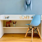 Ein KraftKids Kinderschreibtisch weiß mit Zwischenboden, darauf Holzspielzeug und Buntstifte, dazu ein blauer Stuhl mit Holzbeinen vor einer blauen Wand mit Plüschelefant-die perfekte moderne Kinder-Schreibecke.