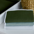 Drei ordentlich gefaltete Handtücher in Dunkelgrün, Hellblau und Weiß liegen gestapelt neben einem geflochtenen Korb und einer passenden KraftKids Wickelunterlage Doppelkrepp Grün Herbstgrün im Hintergrund.