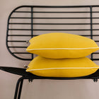 Zwei Kissenbezüge Doppelkrepp Gelb Mustard von KraftKids mit weißer Paspel sind auf einem modernen schwarzen Metallstuhl mit Lattenrost vor einer neutralen beigen Wand gestapelt.