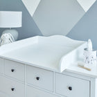 Ein Wickelaufsatz Universal weiß durchgehender Boden von KraftKids mit schwarzen runden Knöpfen steht vor einer grauen und weißen geometrischen Wand. Obenauf stehen eine Lampe mit einem ausgestopften Hasen, eine Taschentuchbox und eine Babyhaarbürste.